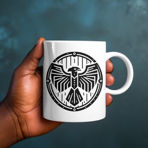 May include: A white ceramic mug featuring a black graphic print of a stylized eagle with spread wings enclosed within a circular border. The design consists of geometric lines and shapes, centered on the front of the smooth, cylindrical vessel.