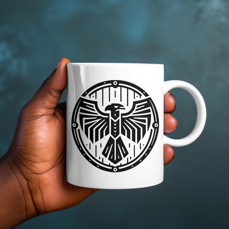 May include: A white ceramic mug featuring a black graphic print of a stylized eagle with spread wings enclosed within a circular border. The design consists of geometric lines and shapes, centered on the front of the smooth, cylindrical vessel.
