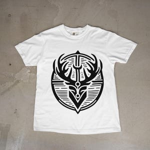 May include: A white short-sleeve t-shirt featuring a large, centered black graphic print. The design depicts a stylized deer head with prominent antlers enclosed within a circular frame composed of horizontal lines and geometric shapes.
