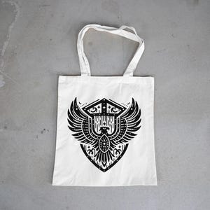 May include: A white tote bag featuring a black graphic print of a stylized eagle with spread wings centered within a shield-like emblem. The design incorporates geometric lines, dots, and symmetrical patterns, with two fabric handles at the top.
