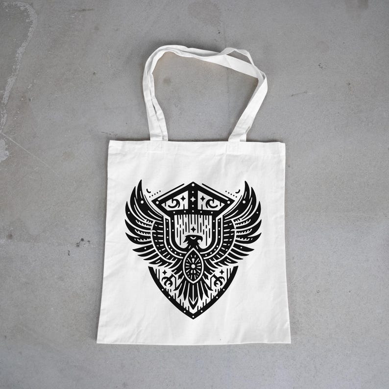 May include: A white tote bag featuring a black graphic print of a stylized eagle with spread wings centered within a shield-like emblem. The design incorporates geometric lines, dots, and symmetrical patterns, with two fabric handles at the top.