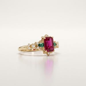 Pode incluir: Um anel de ouro com uma pedra preciosa rosa central de corte esmeralda, ladeada por duas pedras verdes em forma de pera. A banda é decorada com pequenas pedras brancas redondas cravadas numa estrutura de metal detalhada nas laterais.