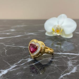 Può includere: Un anello d'oro con una pietra centrale a forma di cuore, un rubino, circondato da diamanti taglio baguette. L'anello presenta un design dettagliato e ornato, su sfondo scuro con un'orchidea bianca.