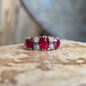 May include: A gold ring featuring three oval-cut rubies and four smaller round diamonds. The rubies are a vibrant red, and the diamonds sparkle against the gold setting. The ring is set on a light-colored surface.