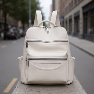 May include: Cream-colored backpack with multiple zippered compartments and side pockets. The bag has a top handle and adjustable shoulder straps. The zippers and hardware are silver-toned. The backpack is made of a smooth, possibly leather-like material.