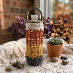 Può includere: Una bottiglia d'acqua in plastica trasparente racchiusa in una custodia all'uncinetto multicolore. La custodia presenta fasce orizzontali di filato verde oliva, crema, arancione bruciato, giallo senape e grigio antracite con un manico.