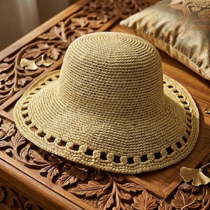 May include: A light tan crochet hat with a rounded crown and a wide brim. The brim features a decorative openwork pattern consisting of a series of small, evenly spaced rectangular cutouts encircling the outer edge of the hat's structure.