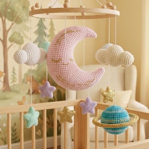 Può includere: Mobile per neonati fatto a mano con una luna a mezzaluna rosa all'uncinetto, dettagli dorati, nuvole bianche e stelle colorate. Un pianeta blu con un anello dorato ed elementi in legno completano il design. Il mobile è appeso sopra una culla di legno.