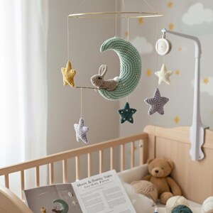 May include: A handmade baby mobile featuring a crocheted green moon with a small bunny, surrounded by yellow, purple, and blue stars. The mobile hangs above a wooden crib, with a teddy bear and craft supplies visible.