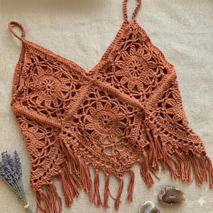 Puede incluir: Un top de ganchillo hecho a mano de color óxido, con un diseño de cuello en V y flecos. El top presenta un patrón floral y tirantes finos. La prenda se exhibe sobre una superficie neutra, mostrando su intrincado diseño calado.