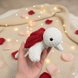 May include: A handmade crochet turtle with a white body and a red shell. The turtle has a black eye and is set against a background of rose petals and fairy lights. This soft toy is a charming decorative item.