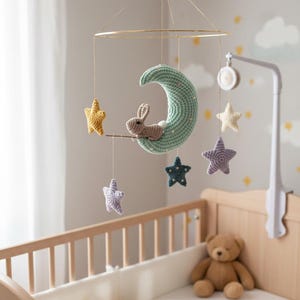 May include: A handmade baby mobile featuring a crocheted crescent moon in light green, a brown bunny, and colorful stars. The mobile hangs above a wooden crib with a teddy bear inside. The nursery decor has a soft, whimsical theme.