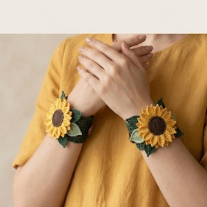 May include: A pair of crochet wrist cuffs featuring large, three-dimensional yellow sunflowers with dark brown centers, accented by small green crocheted leaves. The cuffs are made of a dark green mesh-like crochet pattern and fasten with buttons.