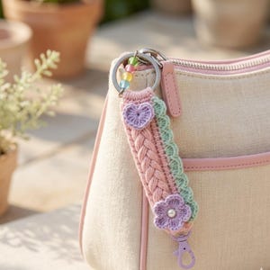 Op de afbeelding: Een handgemaakte sleutelhanger met een gevlochten roze en groene band, versierd met een paars hart en bloem. De sleutelhanger heeft een zilveren ring en een paarse karabijnhaak. Kleurrijke kralen voegen een decoratief tintje toe.