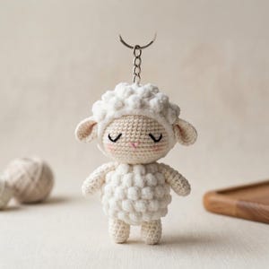 Puede incluir: Un llavero de oveja de ganchillo blanco hecho a mano. La oveja tiene una cabeza esponjosa, orejas pequeñas y un cuerpo texturizado. Presenta ojos cerrados y una boca rosa. El llavero es plateado.