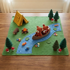 May include: A crochet play mat featuring a green landscape with a blue river, a yellow tent, a brown canoe with a bear and fox, four green pine trees, a campfire, a log, small colorful flowers, mushrooms, and several small fish.