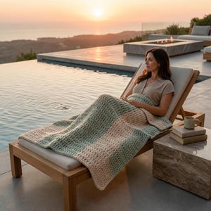 May include: A woman relaxes on a wooden chaise lounge by a pool, covered with a green and beige crocheted blanket. A mug and books sit on a side table. The scene is set at sunset, with the ocean in the background.