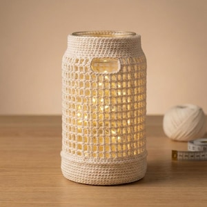 Op de afbeelding: Een crèmekleurige gehaakte lantaarn met een glazen pot erin, verlicht door kleine, warmwitte lampjes. De lantaarn heeft een cilindrische vorm met een handgreepopening en staat op een houten oppervlak. Een bol garen en een meetlint zijn ook zichtbaar.