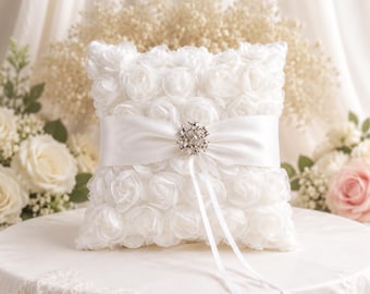 May include: A square white ring pillow covered in fabric roses. A smooth white satin ribbon wraps horizontally around the center, secured by a round, silver-toned brooch featuring clear crystals. Two long, thin white satin ribbons hang down.