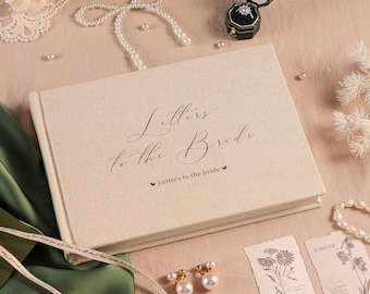 May include: A beige linen-covered book titled "Letters to the Bride" with elegant script. The scene includes a ring box, pearl jewelry, earrings, and floral cards, suggesting a wedding theme.