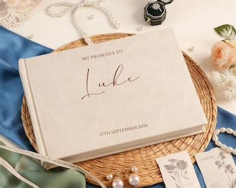 May include: A cream-colored wedding vow book with the inscription "MY PROMISES TO Luke" and the date "17TH SEPTEMBER 2024". The book is on a woven mat, surrounded by pearls, a ring box, and floral accents.