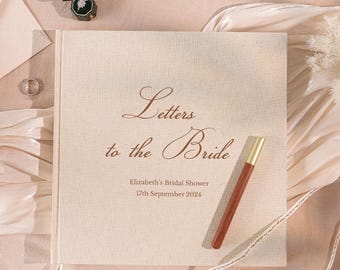 May include: A beige linen-covered guest book with the words "Letters to the Bride" in brown script. The book includes the text "Elizabeth's Bridal Shower 17th September 2024". A pen with a gold tip rests on the book. A ring box and rings are also visible.