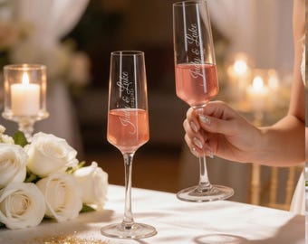 May include: Two champagne flutes filled with a pink beverage. The flutes are engraved with the names "Judy & Luke" and a date. The glasses are on a white table with white roses and a lit candle in the background.