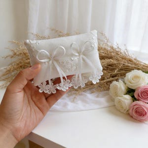 May include: A white rectangular ring pillow with delicate lace trim and two ribbon bows. The pillow is decorated with floral lace and has a soft, textured appearance. The pillow is held in a hand, with a bouquet of roses in the background.