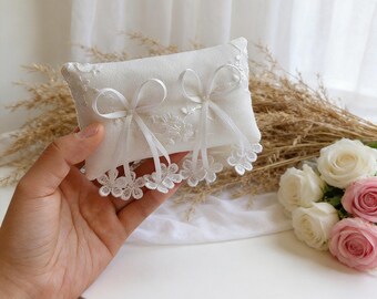 May include: A white rectangular ring pillow with delicate lace trim and two ribbon bows. The pillow is decorated with floral lace and has a soft, textured appearance. The pillow is held in a hand, with a bouquet of roses in the background.
