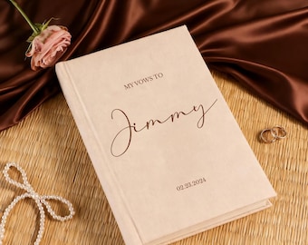 May include: A cream-colored hardcover vow book with dark brown text that reads MY VOWS TO Jimmy and the date 02.23.2024. The book features a fabric-textured cover and a matching ribbon bookmark extending from the bottom edge.