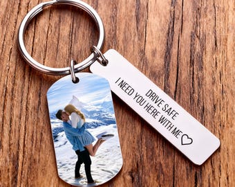 May include: A silver metal keychain with two attached tags. One tag has a photo of a couple kissing in front of a snowy mountain backdrop. The other tag has the engraved text: "DRIVE SAFE I NEED YOU HERE WITH ME" with a heart symbol.