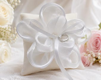 May include: A square off-white ring pillow featuring a large, multi-layered white satin ribbon flower centered on the top. A single silver-toned metal ring is secured by a thin white ribbon loop extending from the center of the fabric flower.