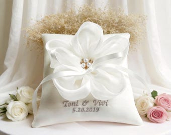 May include: A white satin ring pillow with a large, layered flower and pearl accents. The pillow has a ribbon and the names "Toni & Vivi" with the date "5.20.2019" embroidered on it. Roses in white and pink are in the background.
