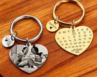 May include: Two personalized keychains on a wooden surface. One keychain has a heart-shaped pendant with a family photo, and the other has a gold heart-shaped pendant with a calendar design. Both keychains have a small round charm with a name and a heart.