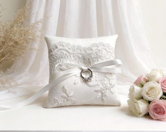 May include: A white ring pillow with intricate lace detailing and a satin ribbon. A silver ring with three stones is secured to the pillow. A bouquet of pink and white roses sits to the side, suggesting a wedding theme.