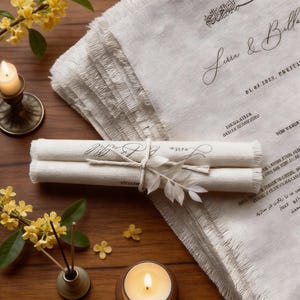 Puede incluir: Una pila de tela color crema con flecos y texto manuscrito. Rollos enrollados atados con hilo y una hoja decorativa en primer plano. Una vela encendida y flores amarillas sobre una superficie de madera.