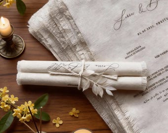 May include: A stack of off-white, fringed fabric with handwritten text. Rolled scrolls tied with twine and a decorative leaf are in the foreground. A lit candle and yellow flowers are on a wooden surface.
