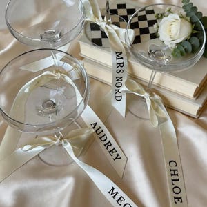 Puede incluir: Cuatro copas de champán transparentes con cintas color marfil atadas alrededor de los tallos. Las cintas están impresas con nombres. Una copa contiene una rosa blanca y vegetación. El fondo incluye libros y una superficie satinada.