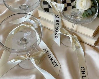 May include: Four clear champagne coupe glasses with ivory ribbons tied around the stems. The ribbons are printed with names. One glass contains a white rose and greenery. The background includes books and a satin surface.