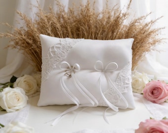 May include: A white satin ring pillow with lace trim and two ribbon bows. Two rings are attached to the pillow with white ribbons. The pillow is surrounded by white and pink roses and dried wheat.
