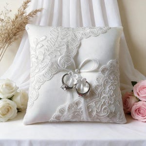 Puede incluir: Un cojín de anillos cuadrado de satén blanco con intrincados detalles de encaje. Dos anillos de plata están sujetos con una cinta blanca y un adorno de perla. El cojín está decorado con encaje floral, un accesorio de boda.