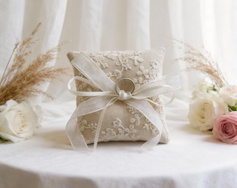 May include: A square, beige ring pillow with floral lace embroidery. A silver ring is attached to the pillow with a white ribbon tied in a bow. The pillow is on a white surface, with flowers and decorative plants in the background.