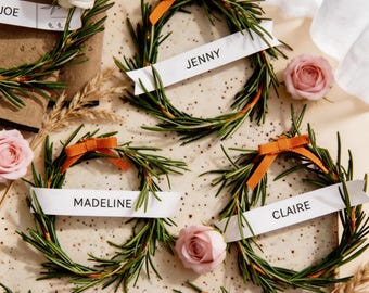 May include: Arrangement of rosemary wreaths with orange bows and white name tags. The tags read "JENNY", "MADELINE", and "CLAIRE". Pink and white roses and wheat stalks are scattered around the wreaths.