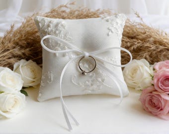 May include: A square, white ring pillow with floral embroidery and a white ribbon tied in a bow. A silver ring is attached to the pillow. White and pink roses and dried plants are in the background.