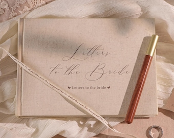 May include: A beige linen-covered book titled "Letters to the Bride" with a matching pen. A white ribbon with text is draped across the book. A silver ring rests on the surface.