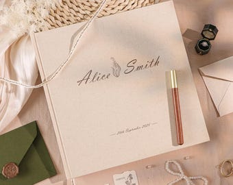 May include: A square, cream-colored photo album with a textured cover featuring the printed text Alice Smith, a small floral icon, and the date 20th September 2025. A wooden pen with a gold tip rests on the cover next to a black ring box.