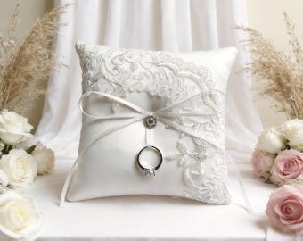 May include: A white satin ring bearer pillow with intricate lace detailing and a ribbon bow. A silver ring with a clear stone is attached to the pillow. The pillow is surrounded by white and pink roses and dried wheat.
