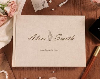 May include: A beige linen-covered wedding guest book with the names "Alice Smith" and the date "20th September 2025" printed in black script. The book is surrounded by pearls, flowers, and stationery.
