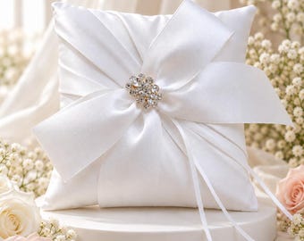 May include: A square white satin ring bearer pillow featuring a large, centered white satin ribbon bow. A cluster of clear, sparkling rhinestones is attached to the center of the bow. Two thin white ribbons hang down from the center of the pillow.