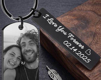 May include: A black keychain with a photo of a smiling couple and a tag that reads "I Love You Forever 02.14.2025." A silver feather charm is also included. The keychain is on a dark surface.